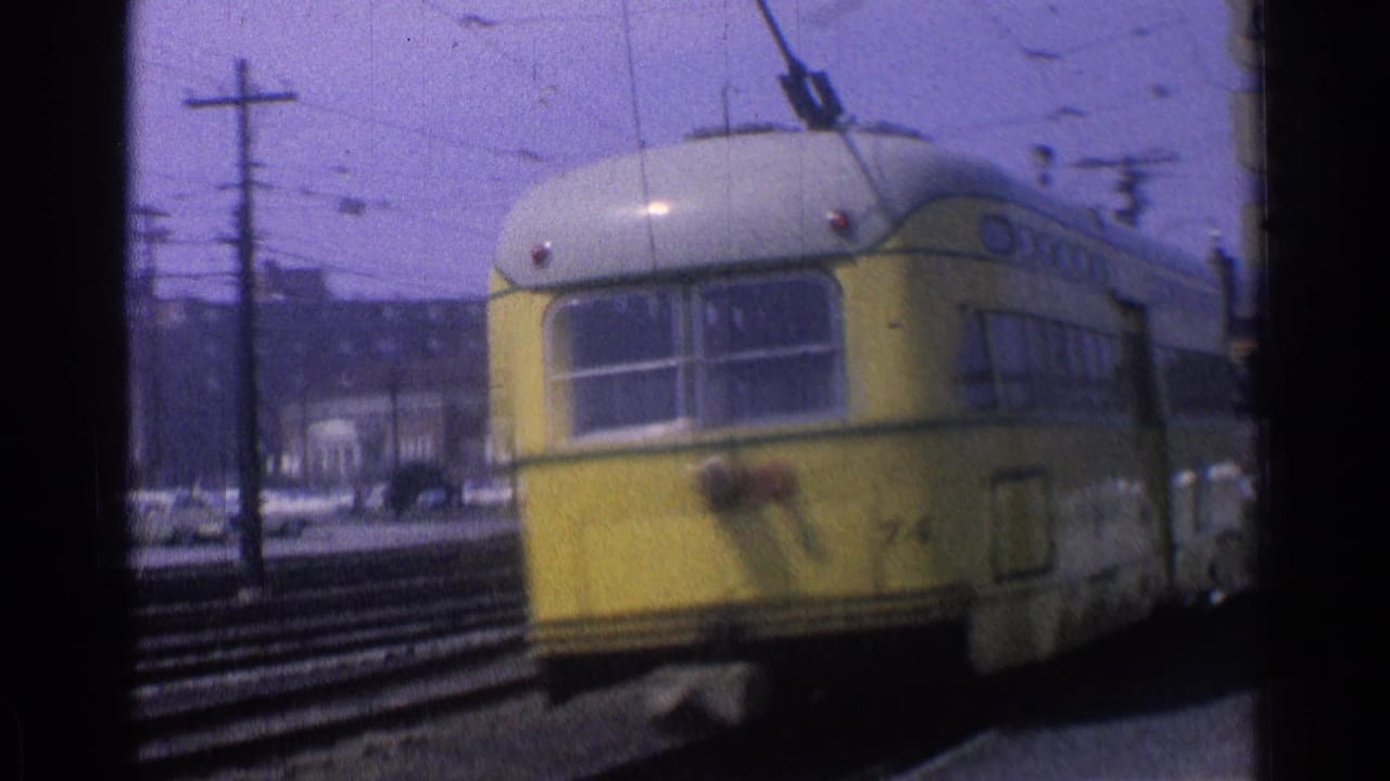 A yellow trolley car is seen from behind on Chicago tracks, with utility poles, wires, and low-rise buildings in the background.