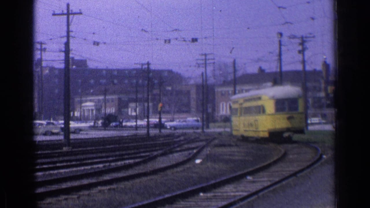 Yellow trolley approaches a curve on rail tracks with utility poles and cars in the background.