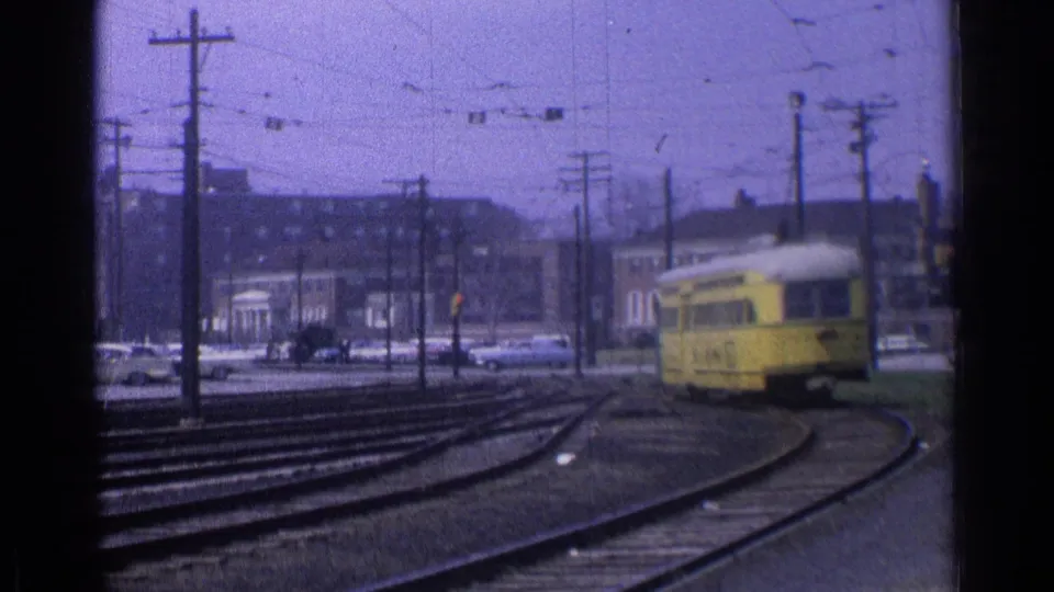 1976 Chicago 8mm frame showing a yellow trolley slowing on curved tracks under overhead wires, with cars and brick buildings beyond.