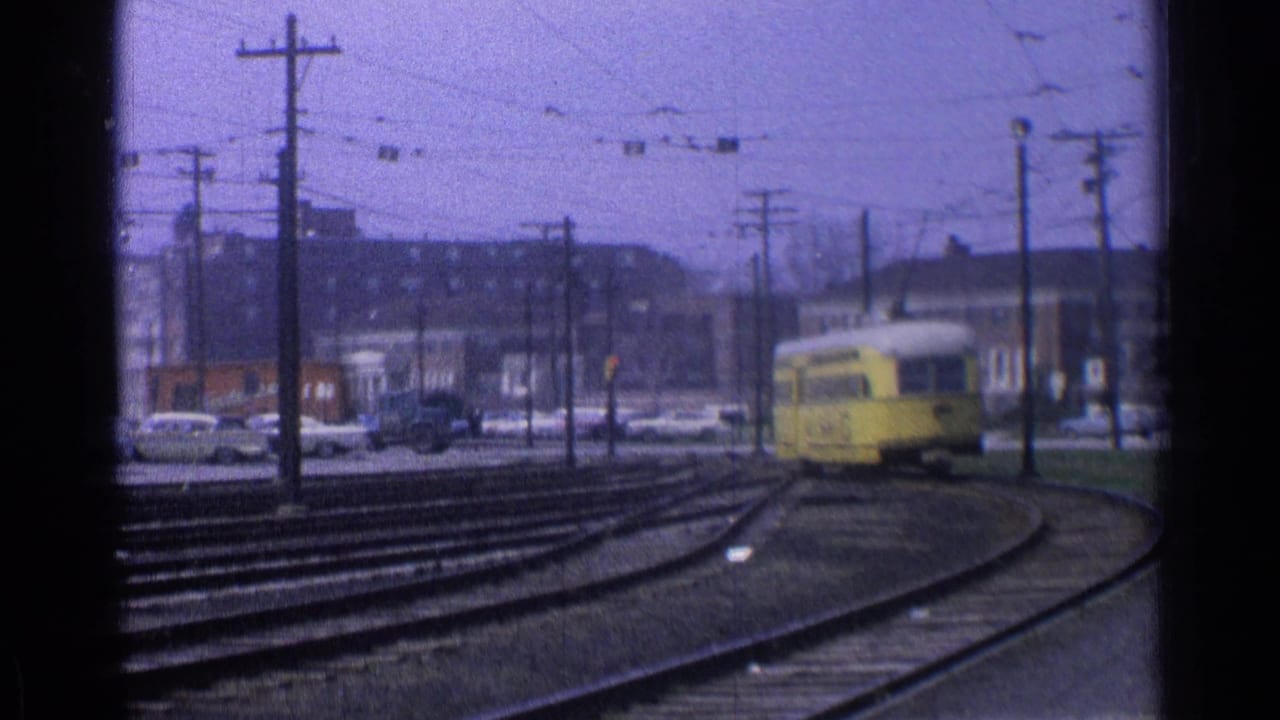 Yellow trolley continues along curved tracks with cars, poles, wires, and buildings behind it.