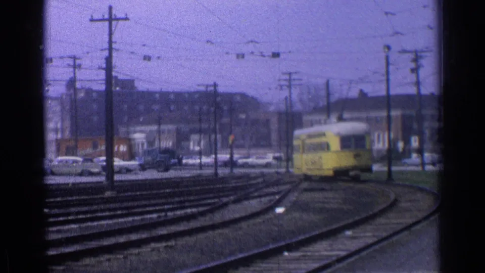 1976 Chicago frame of a yellow trolley on a rail curve, seen through soft 8mm grain with overhead wires and street traffic nearby.