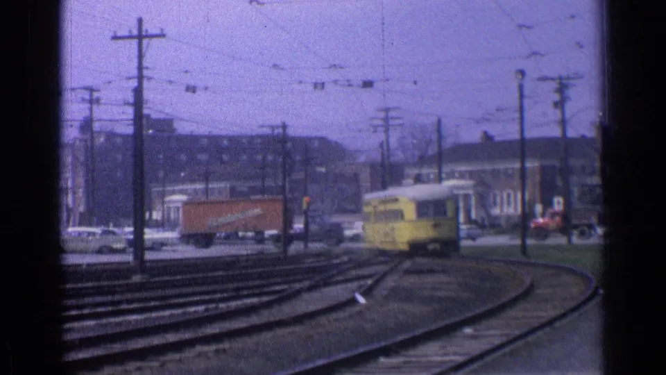 1976 Chicago 8mm frame with a yellow trolley on curved rails, an orange delivery truck, cars, utility poles, and brick buildings.