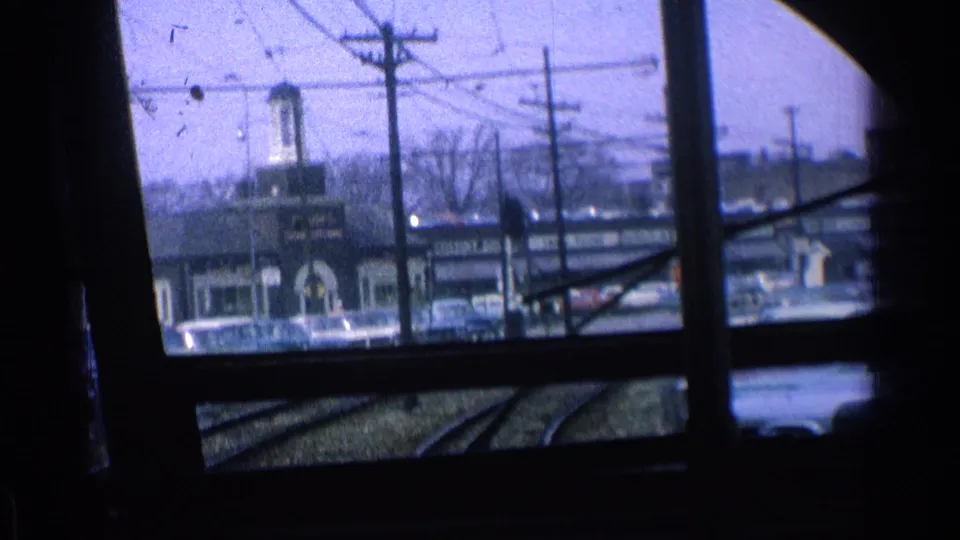 1976 Chicago frame looking through a dark transit or window frame toward rail tracks, utility poles, cars, storefronts, and overhead wires.