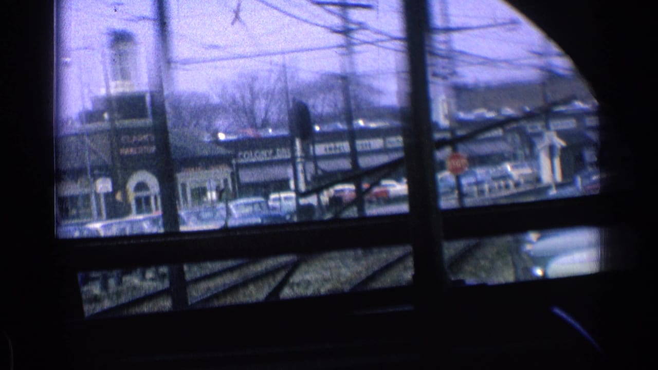 View through train windows toward storefronts, cars, utility lines, and rail tracks.