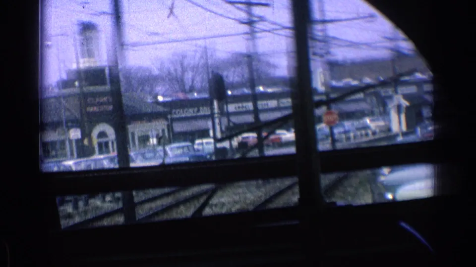 1976 Chicago train-window view with storefronts, vintage cars, utility lines, and rail tracks seen through dark interior framing.