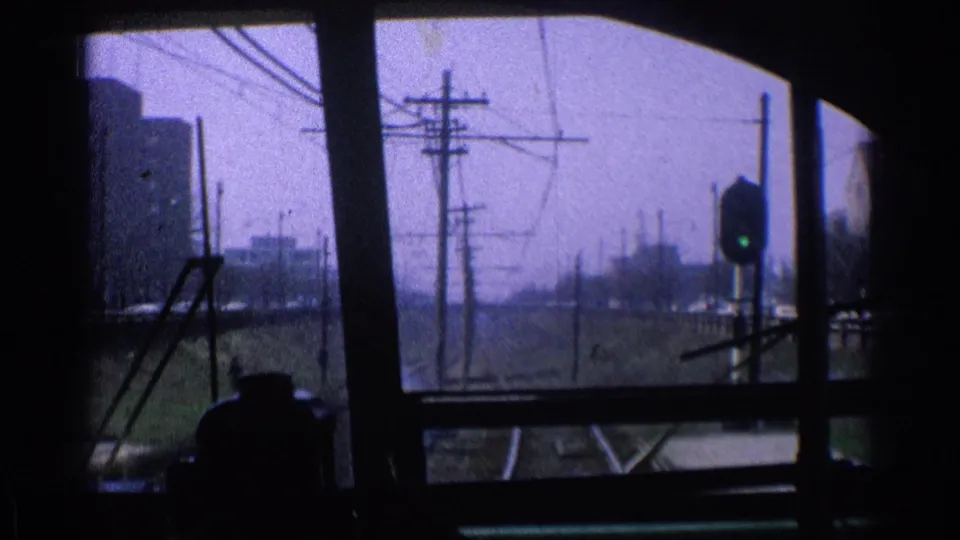 Looking forward from inside the moving train, tracks, utility poles, wires, a signal light, and distant Chicago buildings are visible.