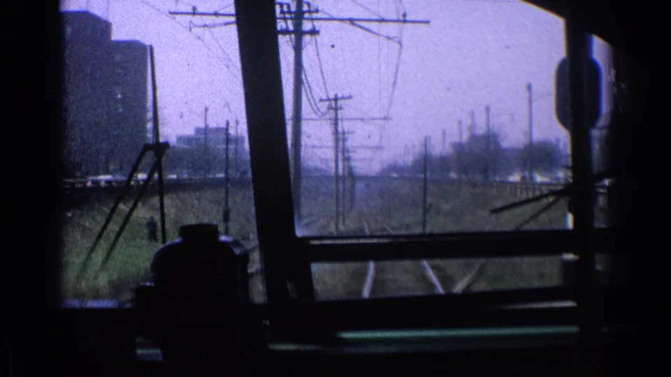A 1976 Chicago rail corridor is seen through the train windows, with tracks, utility poles, wires, and low urban buildings ahead.