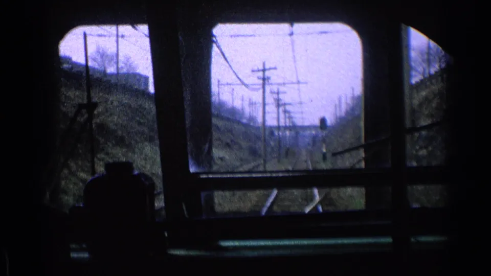 1976 Chicago train-window view showing rails, utility poles, overhead wires, and grassy embankments through dark interior framing.
