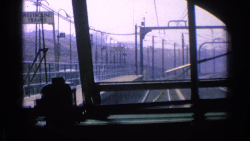 From inside the Chicago transit vehicle, the camera looks along platform railings, tracks, and overhead line supports in hazy light.