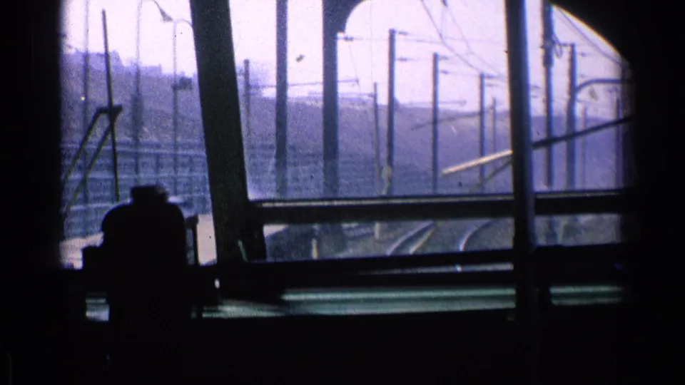 A dim interior frames the 1976 Chicago rail right-of-way, with rails, overhead line poles, fencing, and the operator silhouette visible.