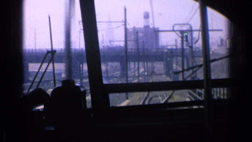 Inside a moving train in 1976 Chicago, silhouetted riders sit before windows overlooking tracks, wires, and industrial structures.