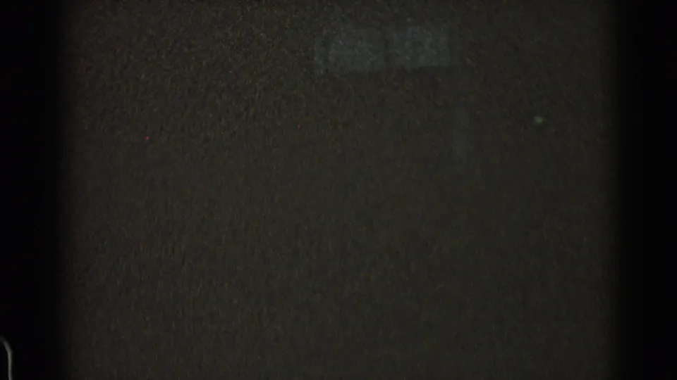 Low-light 8mm frame from the transit sequence, showing a dark wall or tunnel surface with minimal detail and visible film grain.