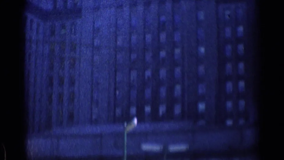 Soft 1976 Chicago street view of a tall windowed building with a streetlamp visible below, tinted blue by aged 8mm film.