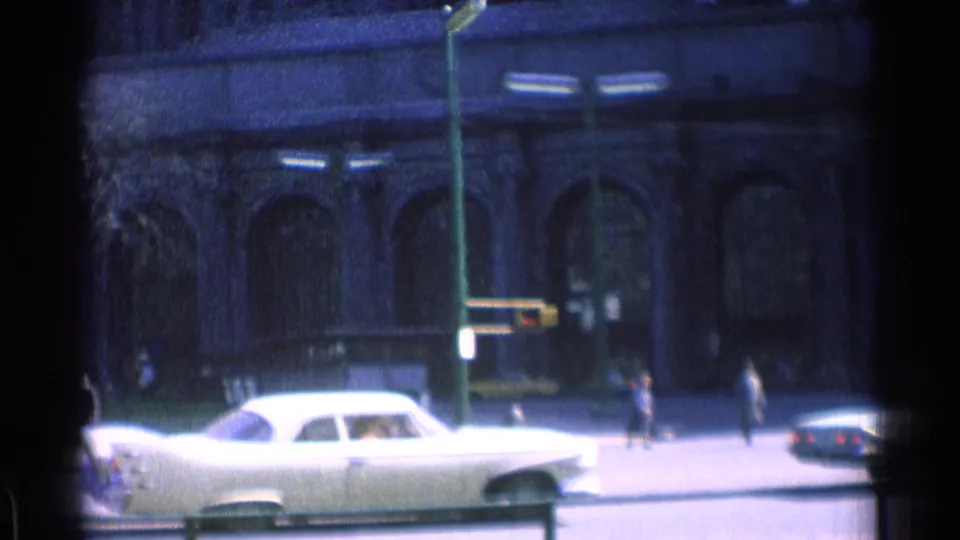 1976 Chicago street frame with a white sedan, pedestrians, streetlamps, and a masonry building with arched ground-floor windows.