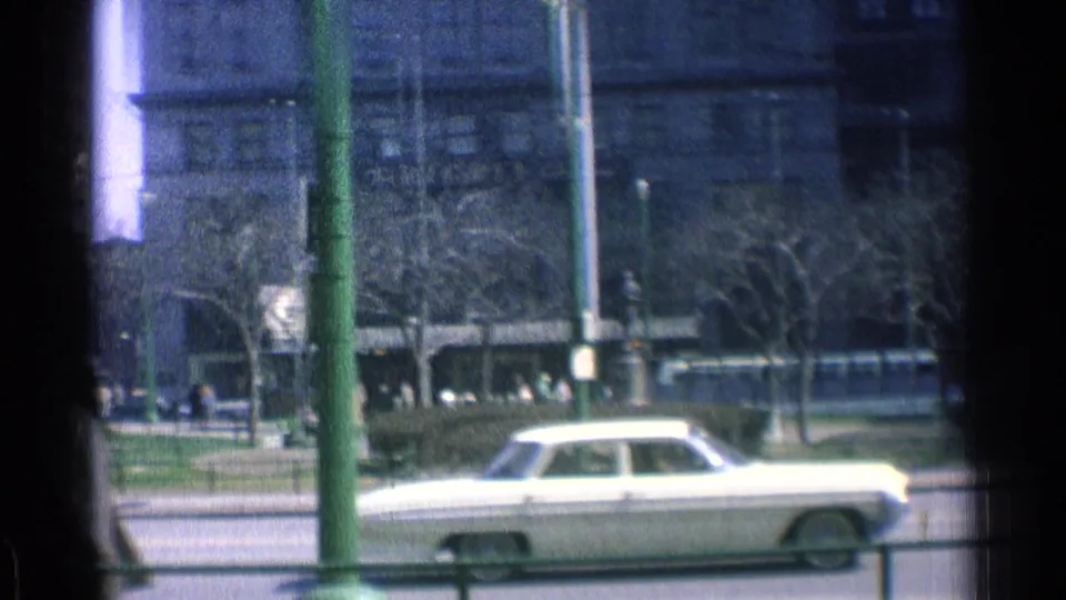 1976 Chicago home-movie frame showing a white sedan on the street below a large windowed building, with poles, bare trees, and pedestrians in view.