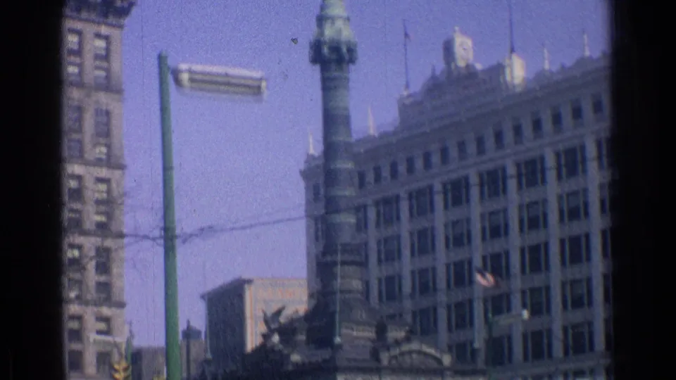 A handheld 8mm view of downtown Chicago buildings, a stone tower structure, a green street pole, overhead wires, and street traffic.