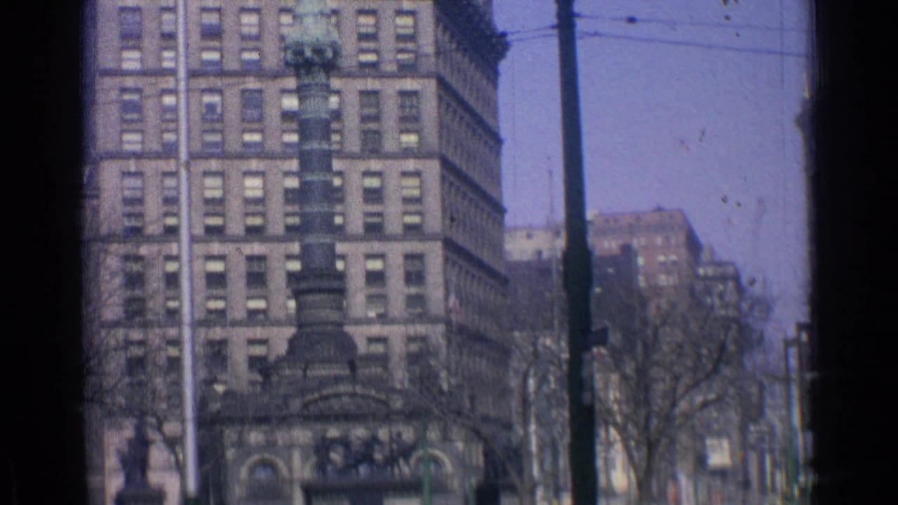 Downtown street view with tall buildings, a dark monument structure, bare trees, and window-frame edges.