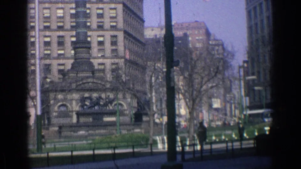 1976 Chicago downtown scene with large masonry buildings, a dark public monument or fountain structure, bare trees, poles, and a low fence.