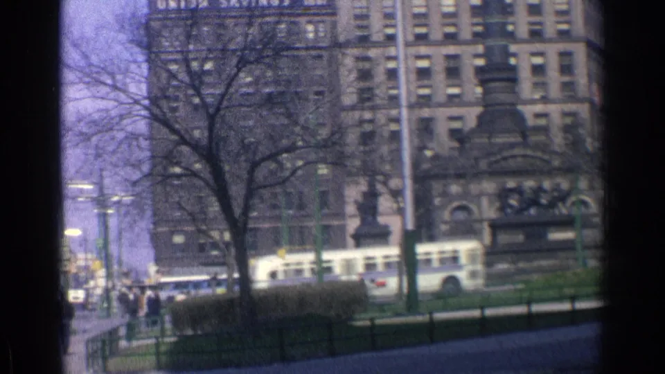1976 Chicago street view with a bus passing near a dark monument structure, tall buildings, bare trees, poles, and small pedestrians.