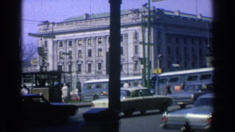 1976 Chicago street scene showing a pale multi-story building, buses, cars, traffic signals, street poles, and pedestrians in the distance.