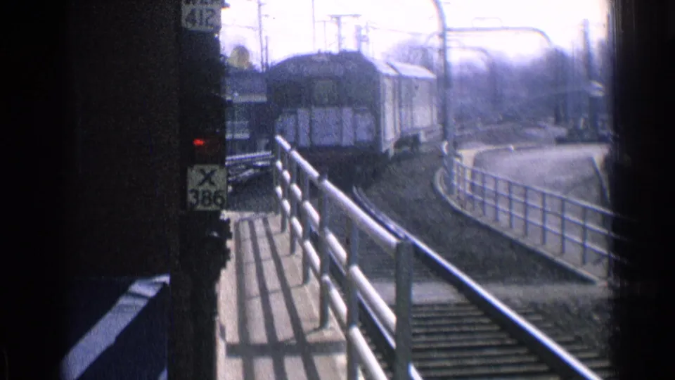 1976 Chicago 8mm frame showing a green passenger train approaching on curved tracks beside a metal platform railing and signal post.