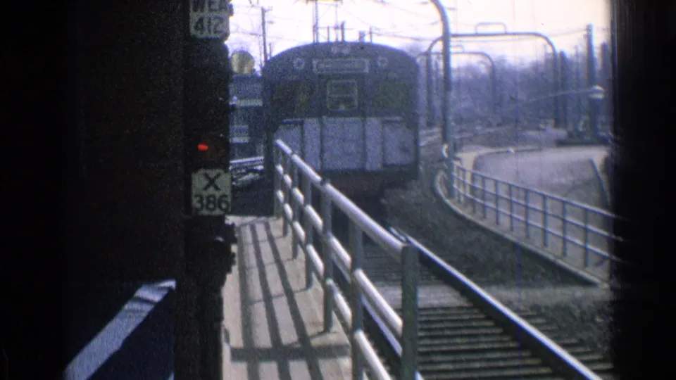 Chicago 1976 home-movie frame of a passenger train approaching the station platform, with railings, signal markings, and overhead lines visible.