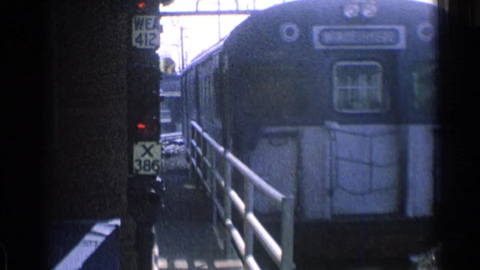 Blue railcar front moves past the Chicago platform viewpoint, with railings, signal signs, and soft 8mm exposure visible.