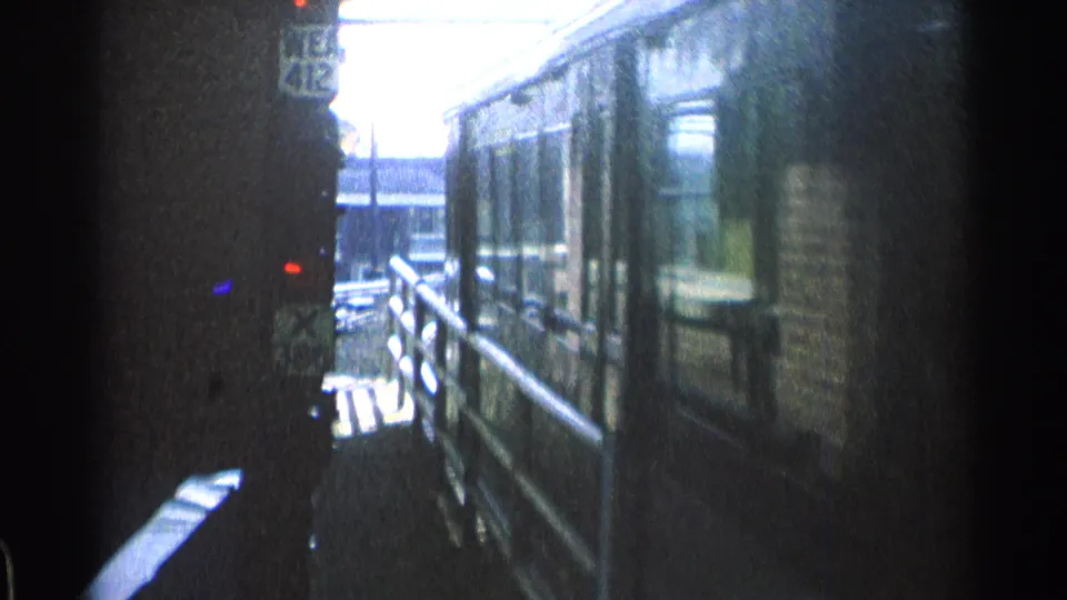 The blue railcar continues past the 1976 Chicago platform viewpoint, with window reflections and station structures beyond the railing.