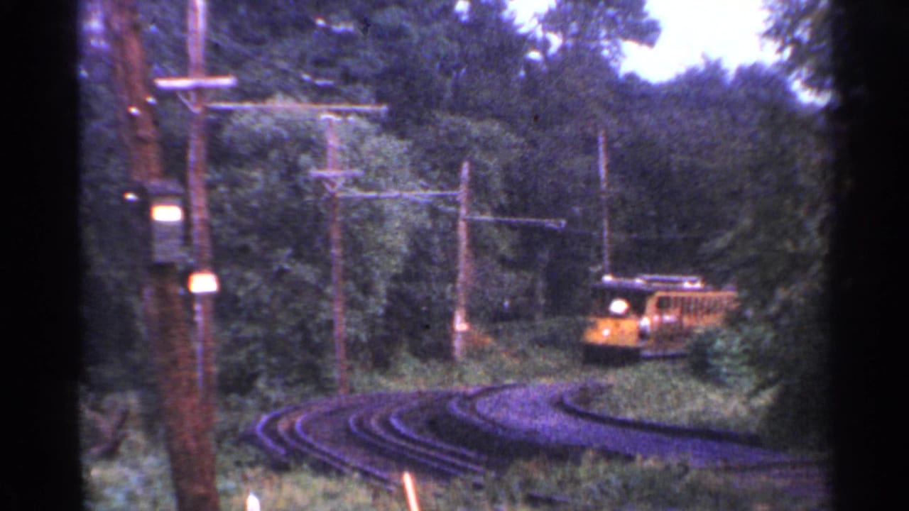 A yellow trolley approaches around a forested rail curve in Connecticut, with overhead wires and utility poles visible.