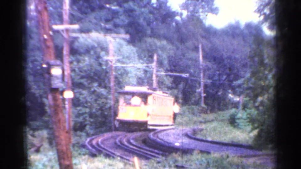 The trolley fills more of the frame as it follows the Connecticut rail curve through dense trees and overhead wiring.