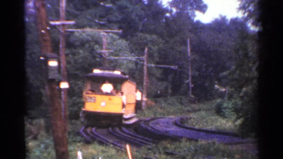 A yellow open-sided trolley nears the camera on a sharp wooded curve, with the front operator area visible in this 1953 Connecticut home movie.