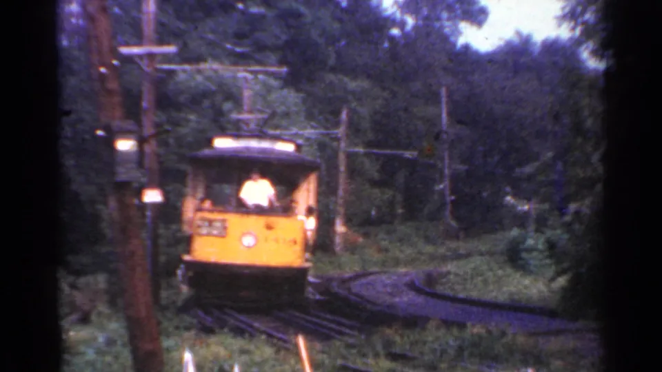 Close 8mm frame of the yellow Connecticut trolley rounding the bend, with a light-shirted person visible at the front and overhead wires behind.
