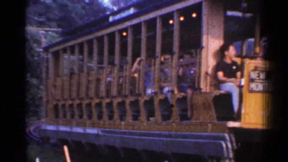 The open-sided yellow trolley passes by on the curved track, with a seated passenger near the front and wooded surroundings behind it.