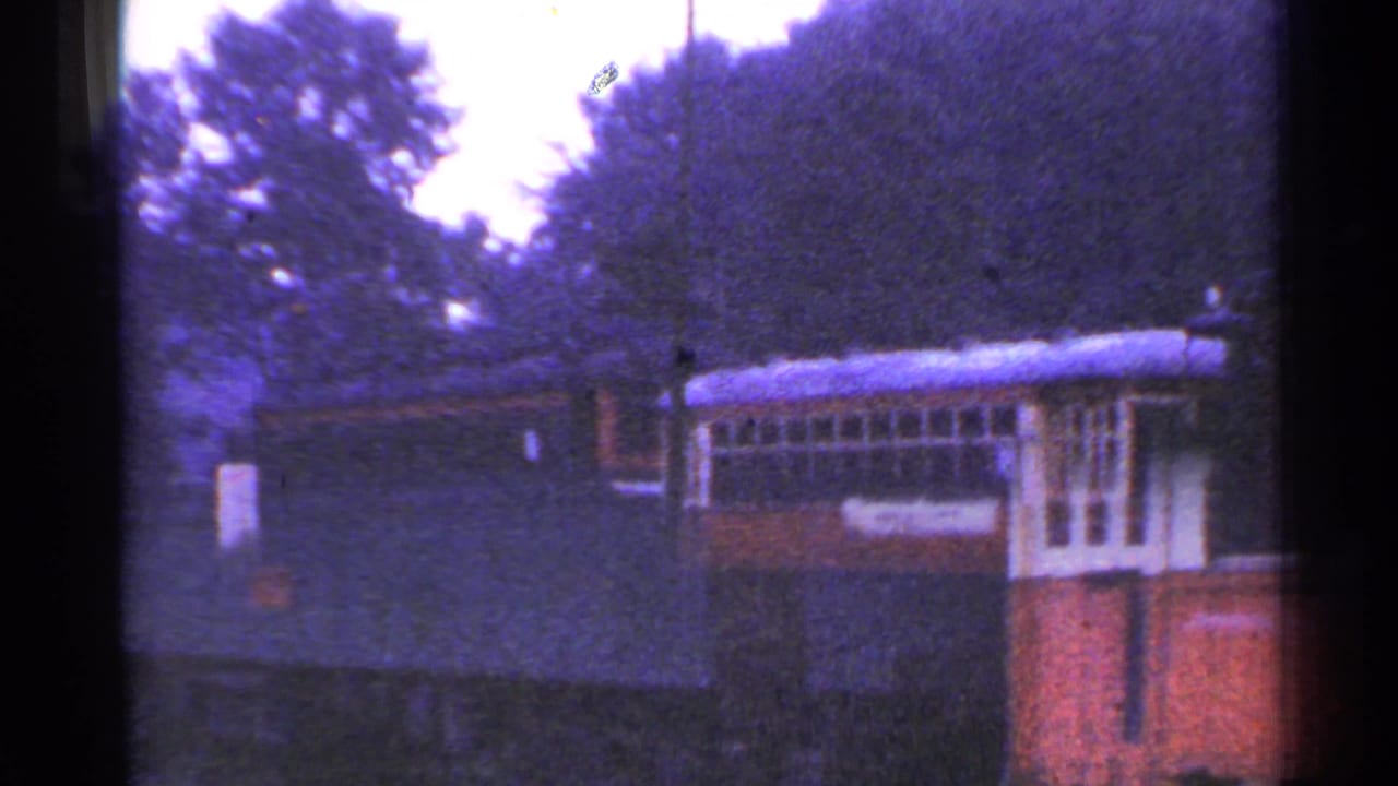 The train car remains beside the station area, with trees, a dark structure, and cloudy daylight in the frame.