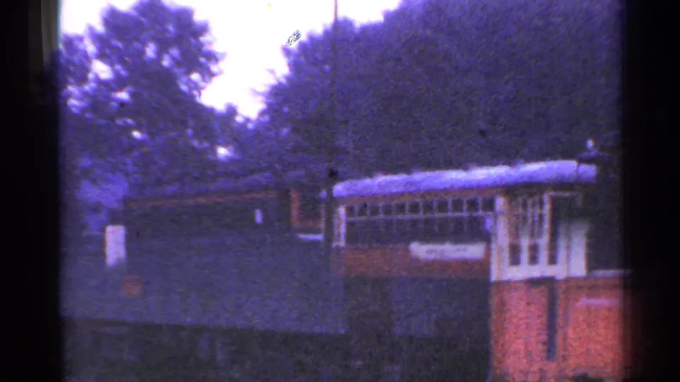 The train car remains beside the station area, with trees, a dark structure, and cloudy daylight in the frame.