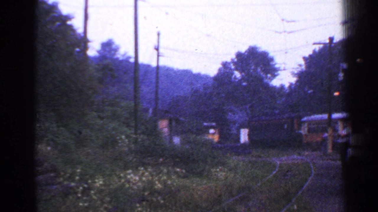 Curving railroad track with trees, utility poles, and train cars in the distance.