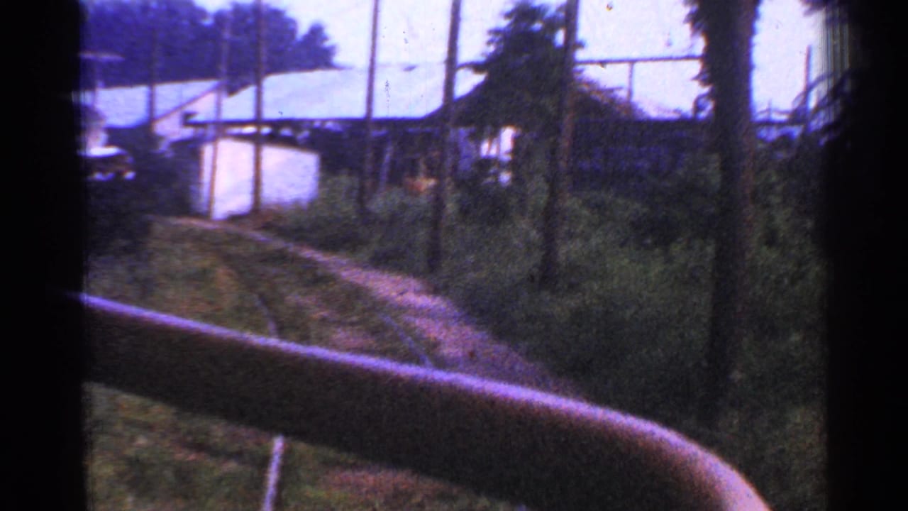 A moving-train view in Connecticut shows curved track, low light-colored buildings, poles, wires, and thick vegetation along the right of way.