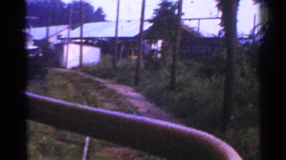 A moving-train view in Connecticut shows curved track, low light-colored buildings, poles, wires, and thick vegetation along the right of way.