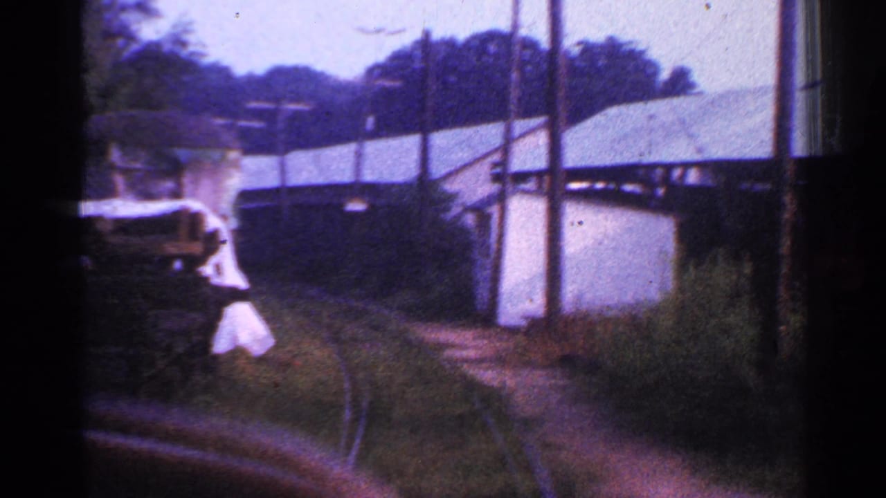 A 1953 Connecticut home-movie frame shows a partially visible adult figure in light clothing near the moving train, with curved track, poles, vegetation, and light-colored buildings beyond.