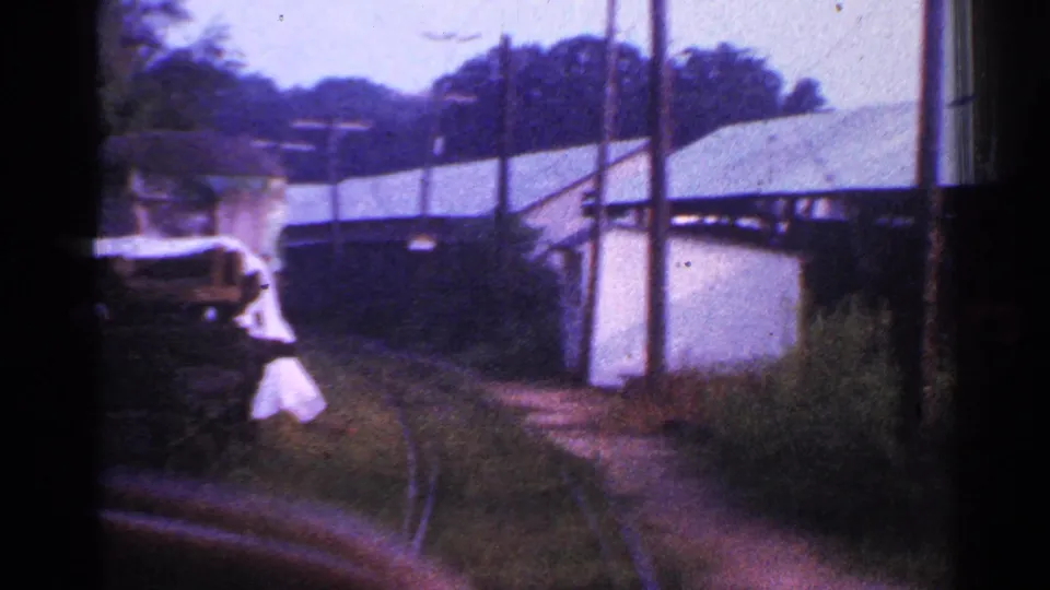 A 1953 Connecticut home-movie frame shows a partially visible adult figure in light clothing near the moving train, with curved track, poles, vegetation, and light-colored buildings beyond.