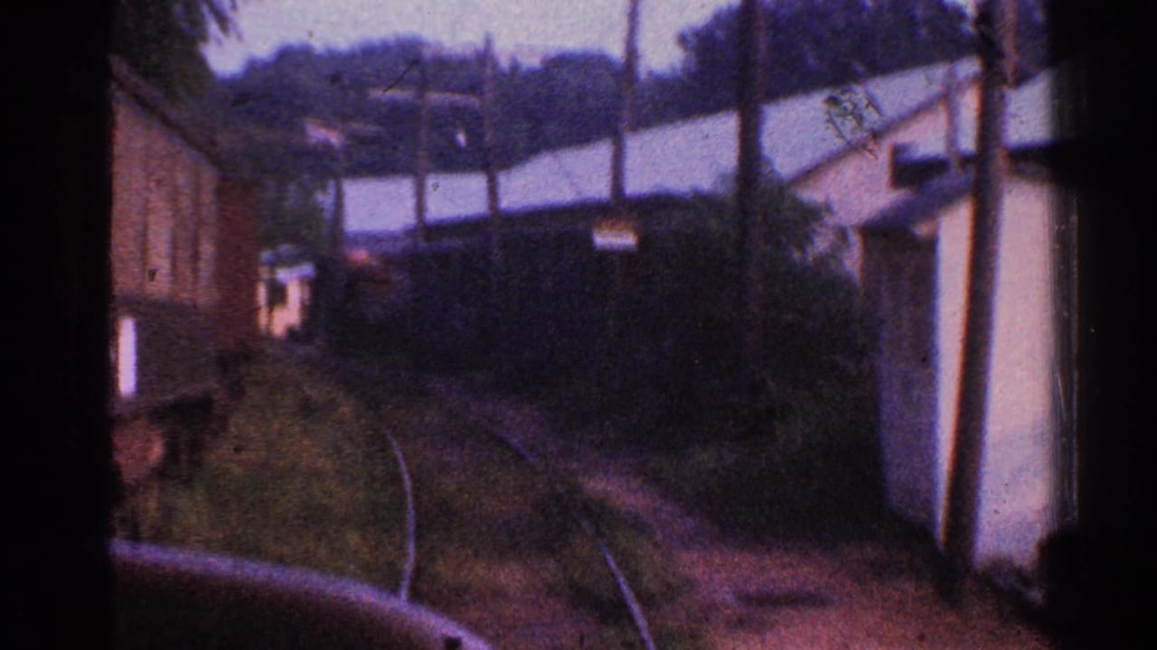 First-person 1953 Connecticut rail view with curving tracks, buildings, and wooded hillside beyond.