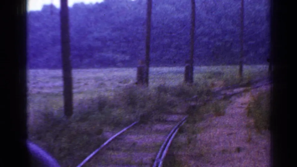 A Connecticut train-window shot shows rails curving beside grass, brush, poles, and a tree-covered hill.