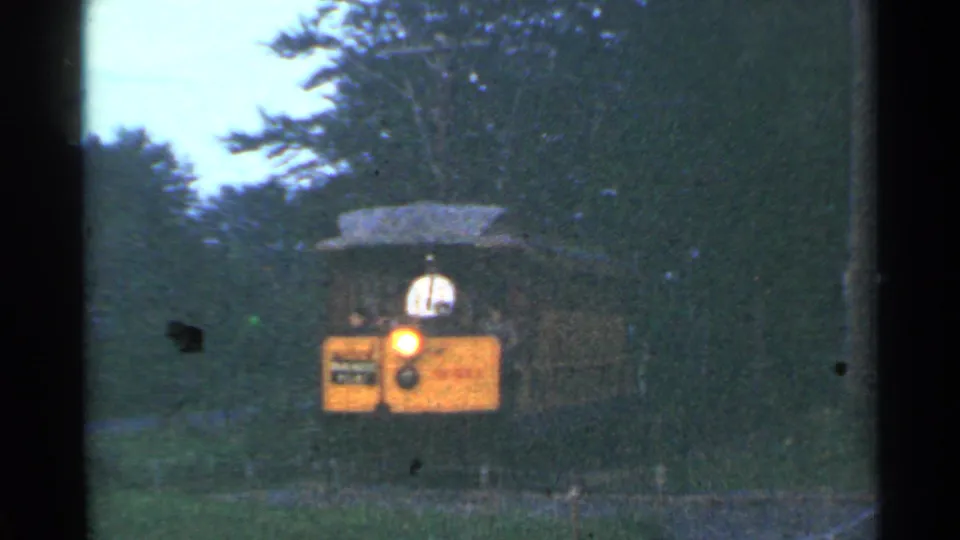 A closer 1953 home-movie view shows the yellow tram front, glowing headlight, driver, and wooded track surroundings.