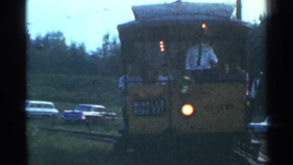 Yellow passenger rail car in Connecticut, 1953, with a glowing headlight, visible riders or crew, parked cars, trees, and station-side track area.