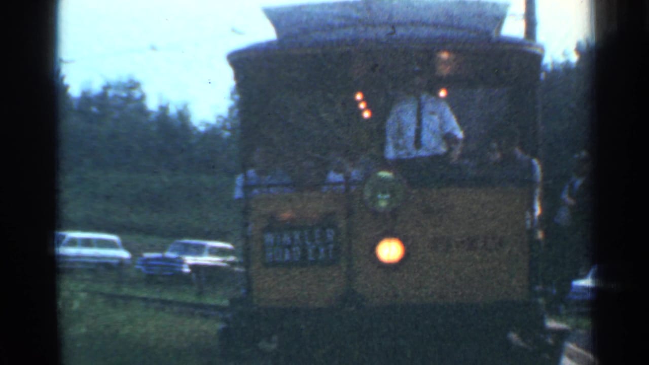1953 Connecticut home-movie frame showing the front of a yellow trolley with a driver, headlamp, roadside greenery, and older cars behind it.