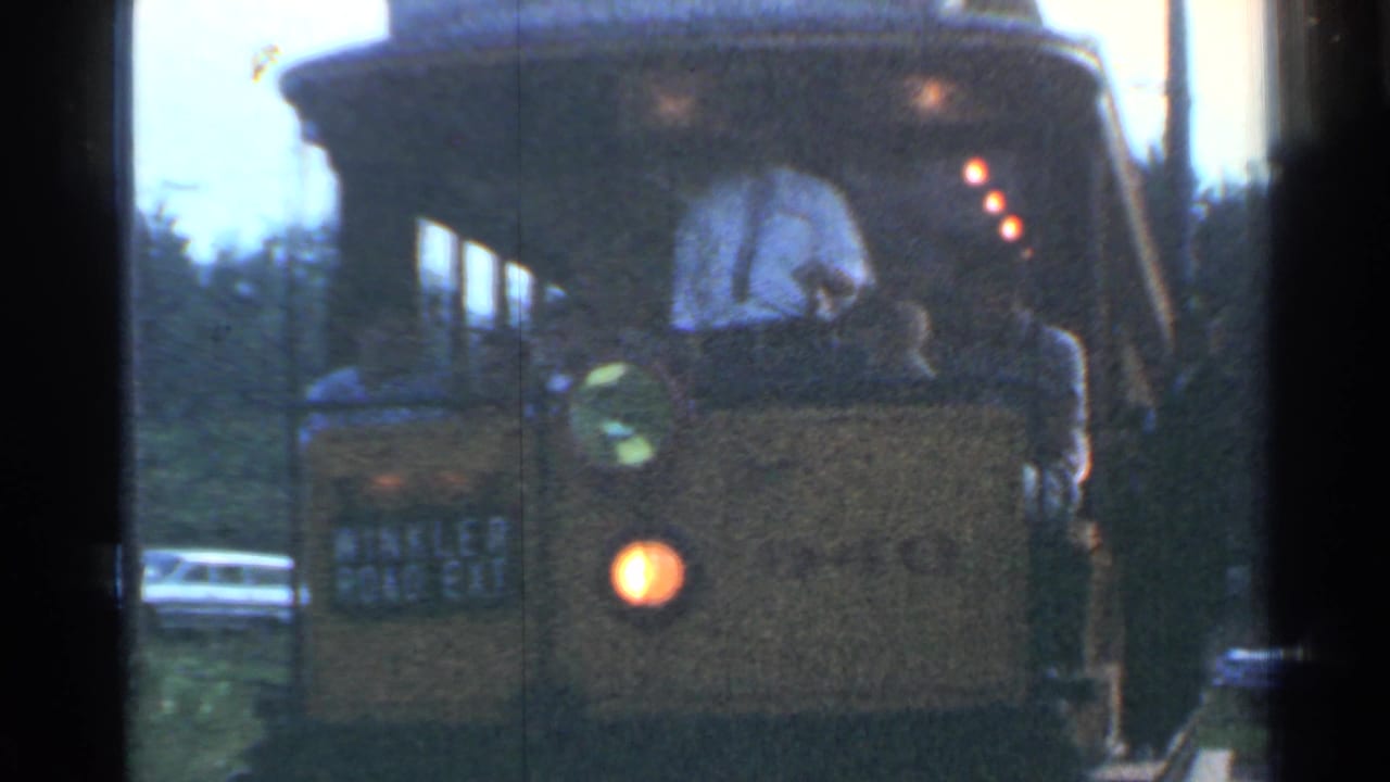 The yellow trolley remains in close view, with the driver at the front, a passenger nearby, and mid-century cars visible along the roadside.