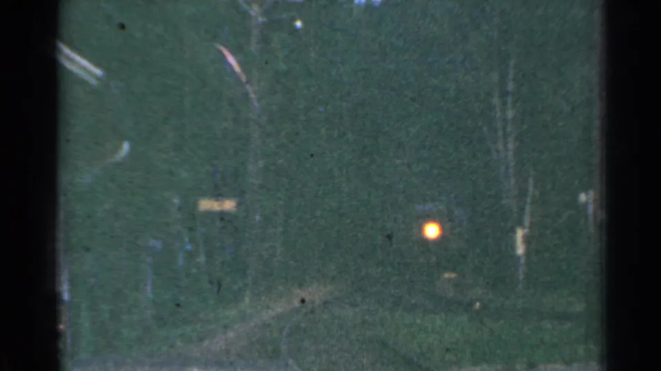 1953 Connecticut 8mm frame looking down a dim rail corridor toward a glowing orange signal light among trees and poles.