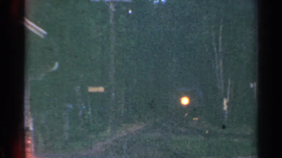 1953 Connecticut home-movie frame showing a low-light rail view with a glowing orange signal, trees, poles and faint overhead wires.