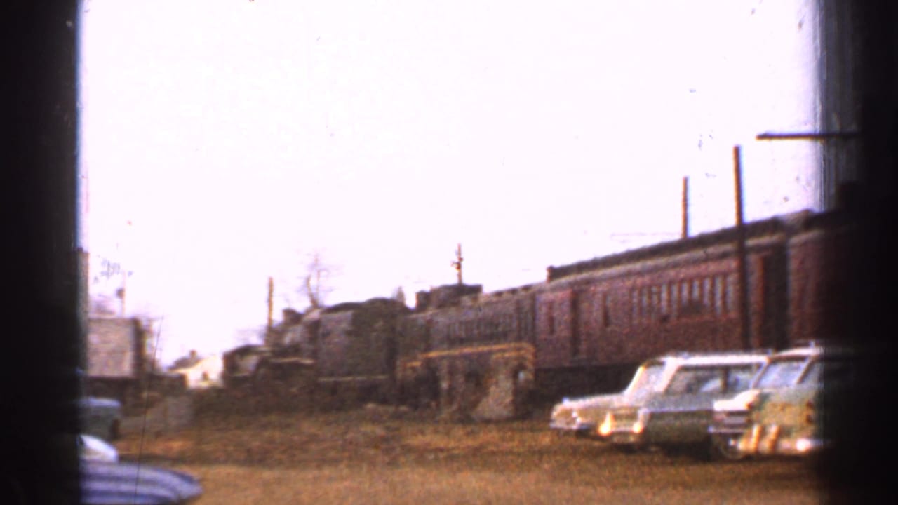 Brown passenger train at a Connecticut station area with 1950s cars parked nearby on a dirt lot.