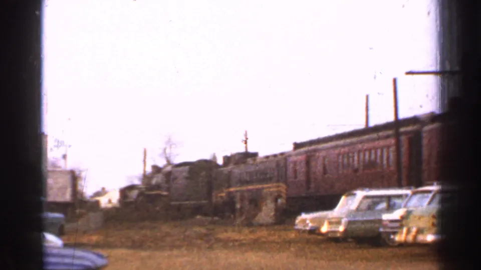 Brown passenger train at a Connecticut station area with 1950s cars parked nearby on a dirt lot.